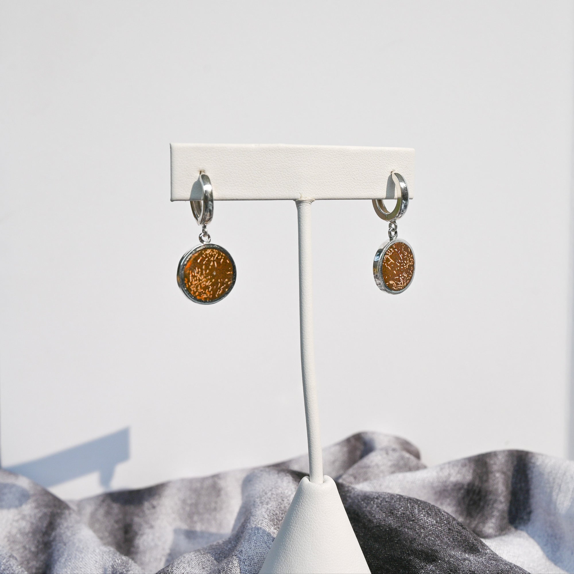 Pair of silver huggie hoop earrings with orange sunburst jasper charms hanging on a white earring stand