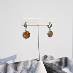 Pair of silver huggie hoop earrings with orange sunburst jasper charms hanging on a white earring stand