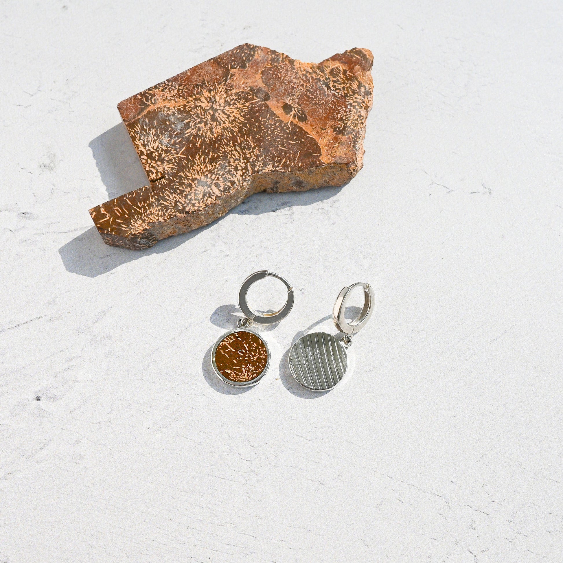 Pair of silver huggie hoop earrings with orange sunburst jasper charms on a textured white surface with a piece of stone.