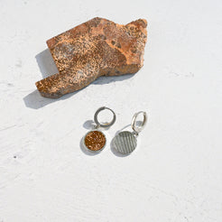 Pair of silver huggie hoop earrings with orange sunburst jasper charms on a textured white surface with a piece of stone.