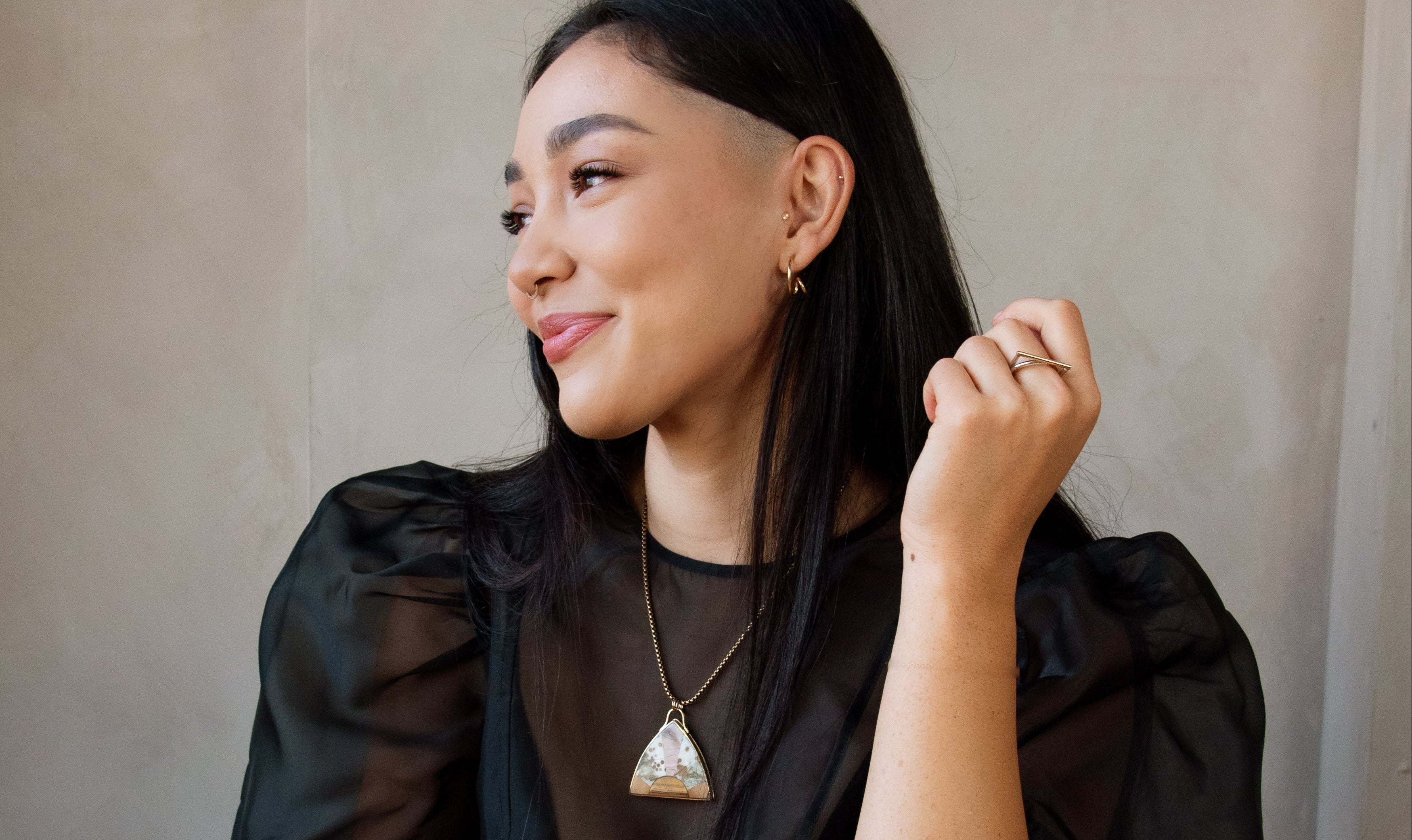 A woman in a black sheer puff-sleeve top , wearing a geometric triangle pendant necklace, in front of a textured beige wall and wooden floor.