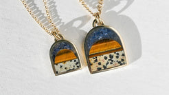Two gold chain necklaces with arched pendants featuring a layered landscape design of blue sky, orange trees, and a speckled beige foreground, displayed on a white textured surface with soft shadows.