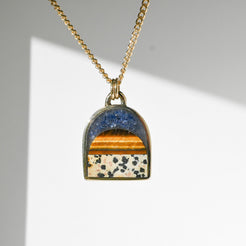 A gold chain necklace with an arched pendant featuring a landscape design with a blue sky, orange and yellow sunset, and a white patterned ground with small black flowers.