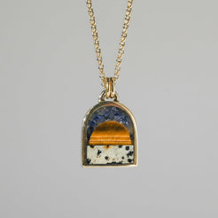 A gold chain necklace with an arched pendant featuring a split design of blue lapis lazuli and tiger's eye on a white speckled background, shown against a white wall with soft shadows.