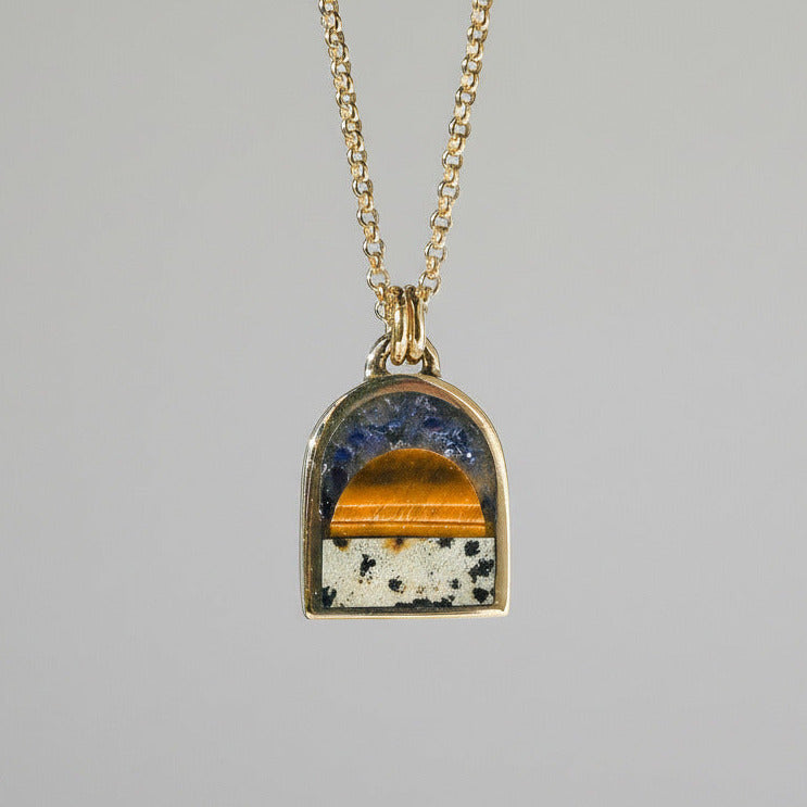 A gold chain necklace with an arched pendant featuring a split design of blue lapis lazuli and tiger's eye on a white speckled background, shown against a white wall with soft shadows.