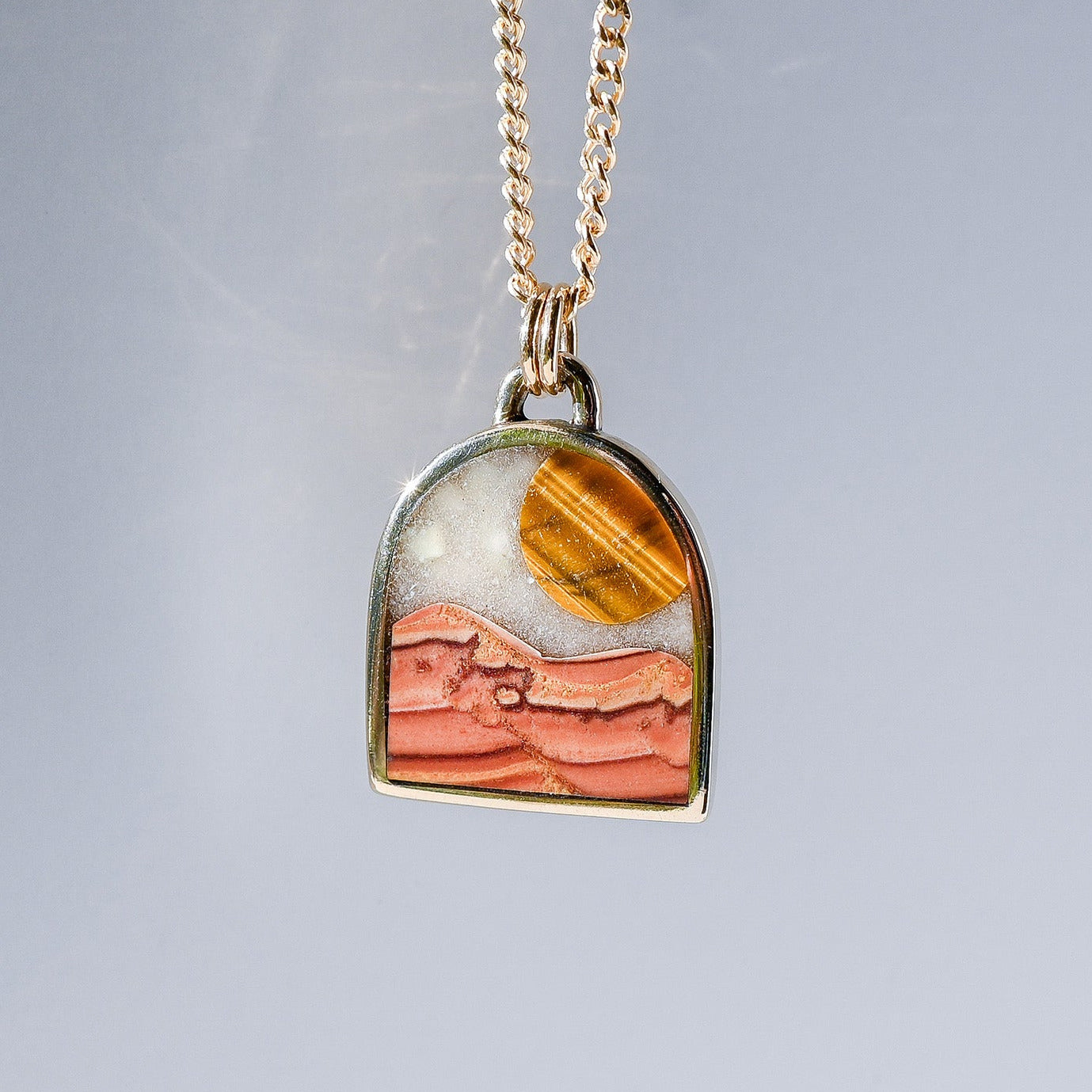 A gold chain necklace with a small arch-shaped pendant featuring a desert landscape design in orange, pink, and white, set against a light gray background.