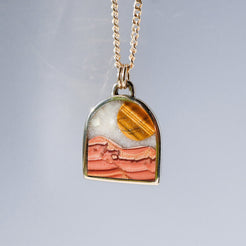 A gold chain necklace with a small arch-shaped pendant featuring a desert landscape design in orange, pink, and white, set against a light gray background.