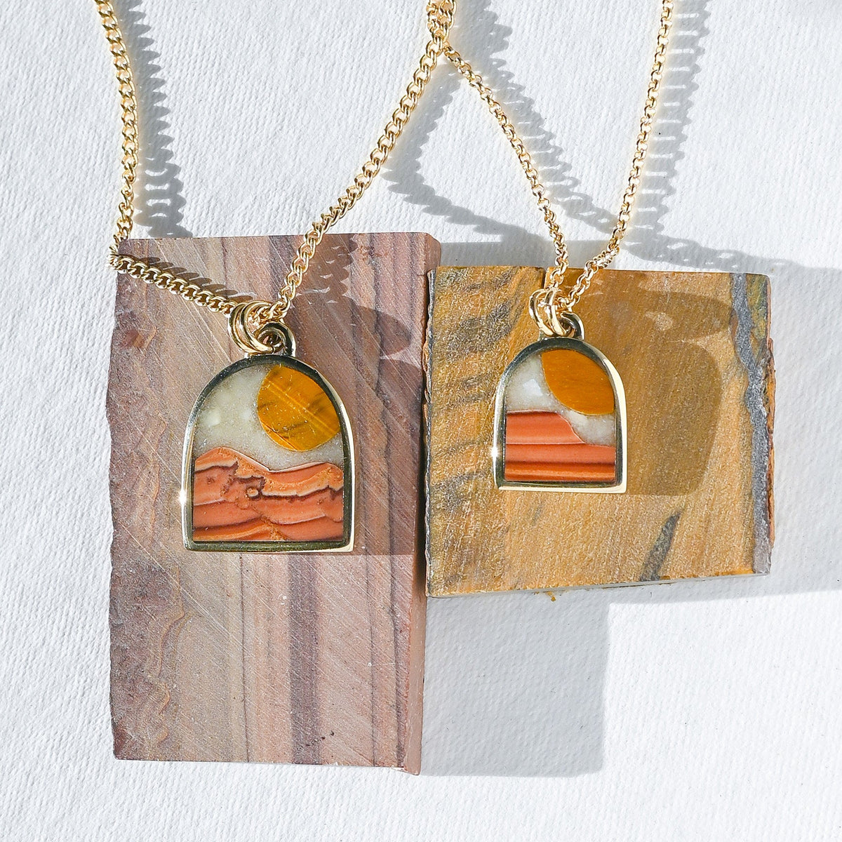 Two gold chain necklaces with arched pendants displayed on rectangular stone slabs. Each pendant features a desert landscape design with orange and white layered rock formations and a yellow sun, set against a white textured background.