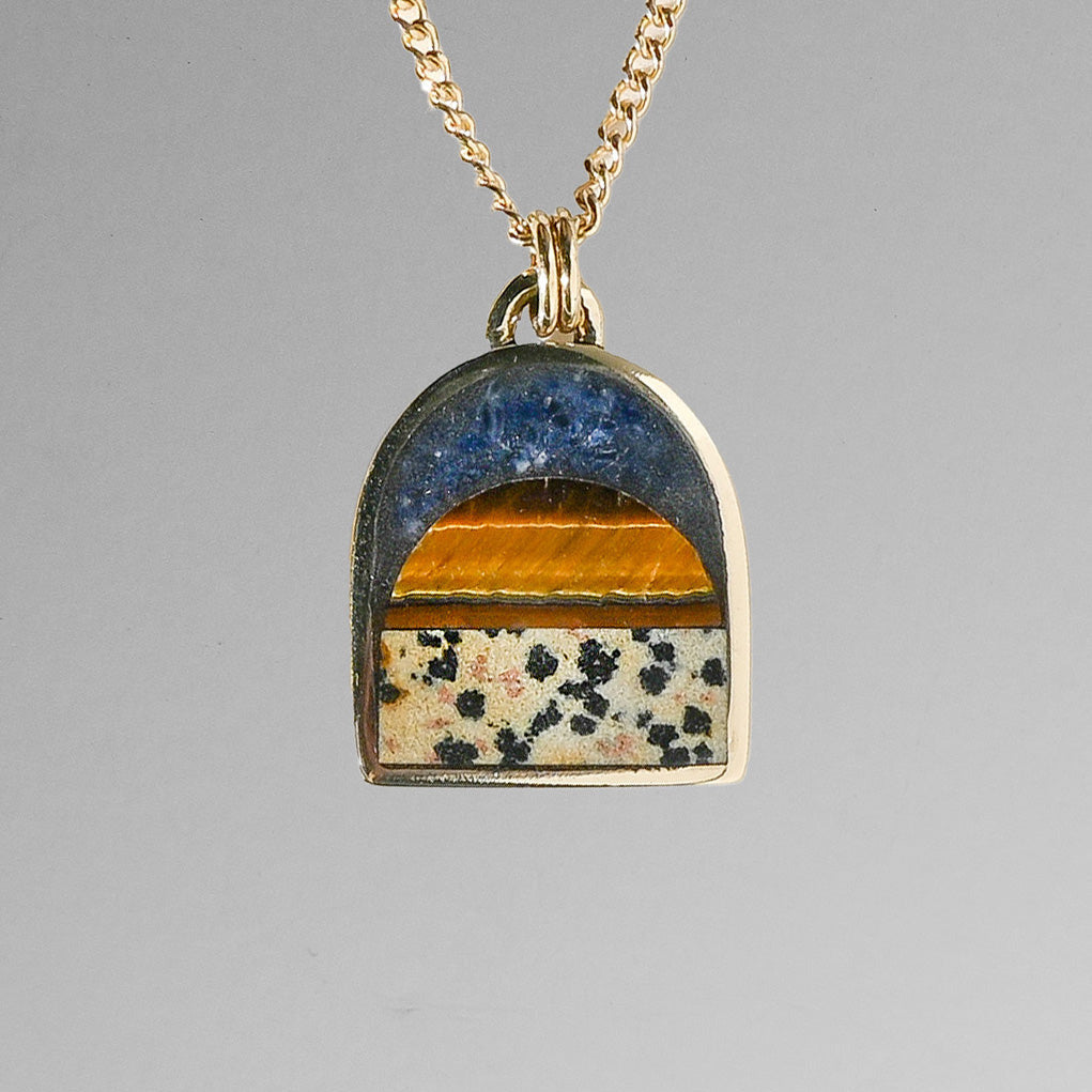 A gold chain necklace with an arched pendant featuring a landscape design with a blue sky, orange sun, and a patterned beige ground with small black flowers.