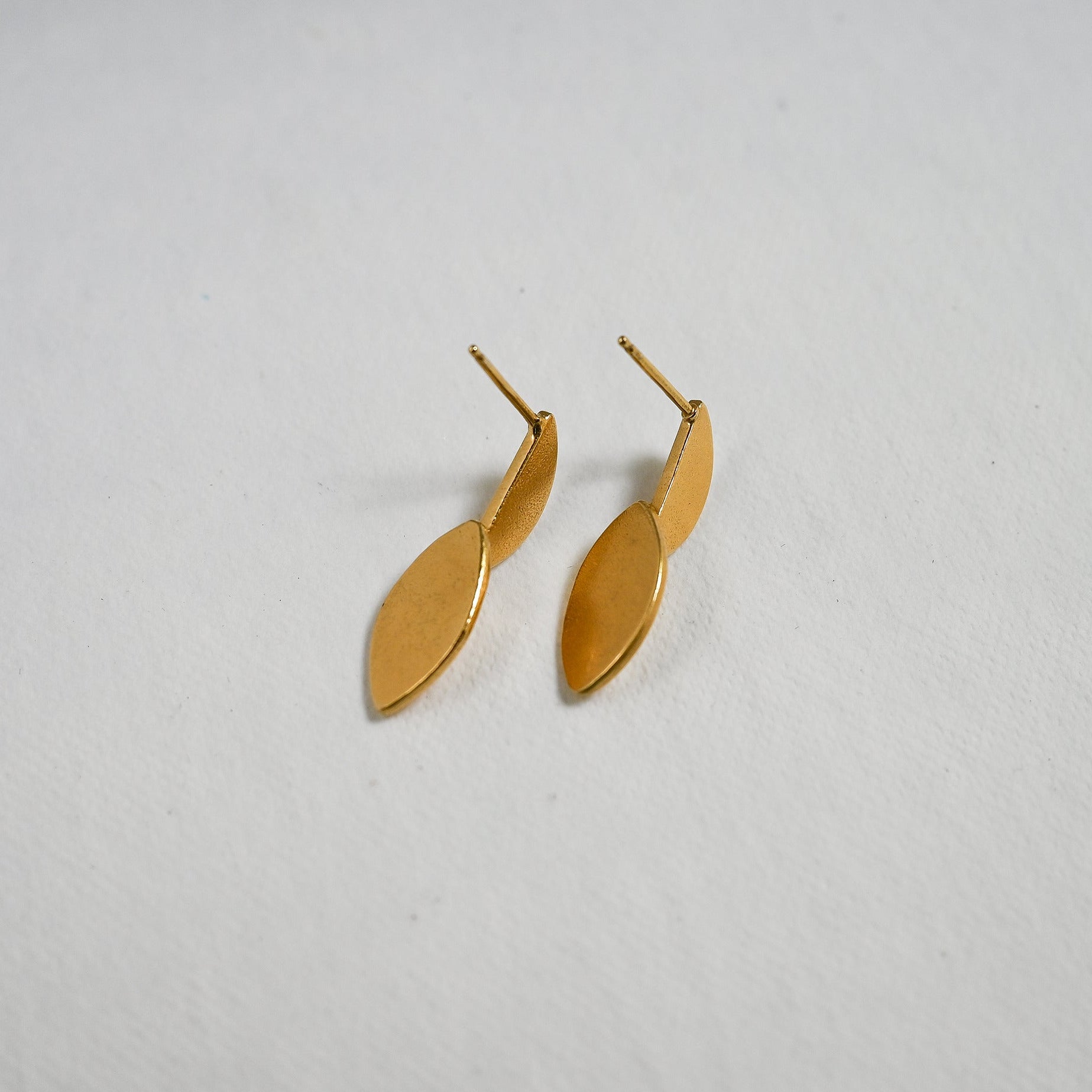 A pair of small gold leaf-shaped dangle earrings on a textured white background.