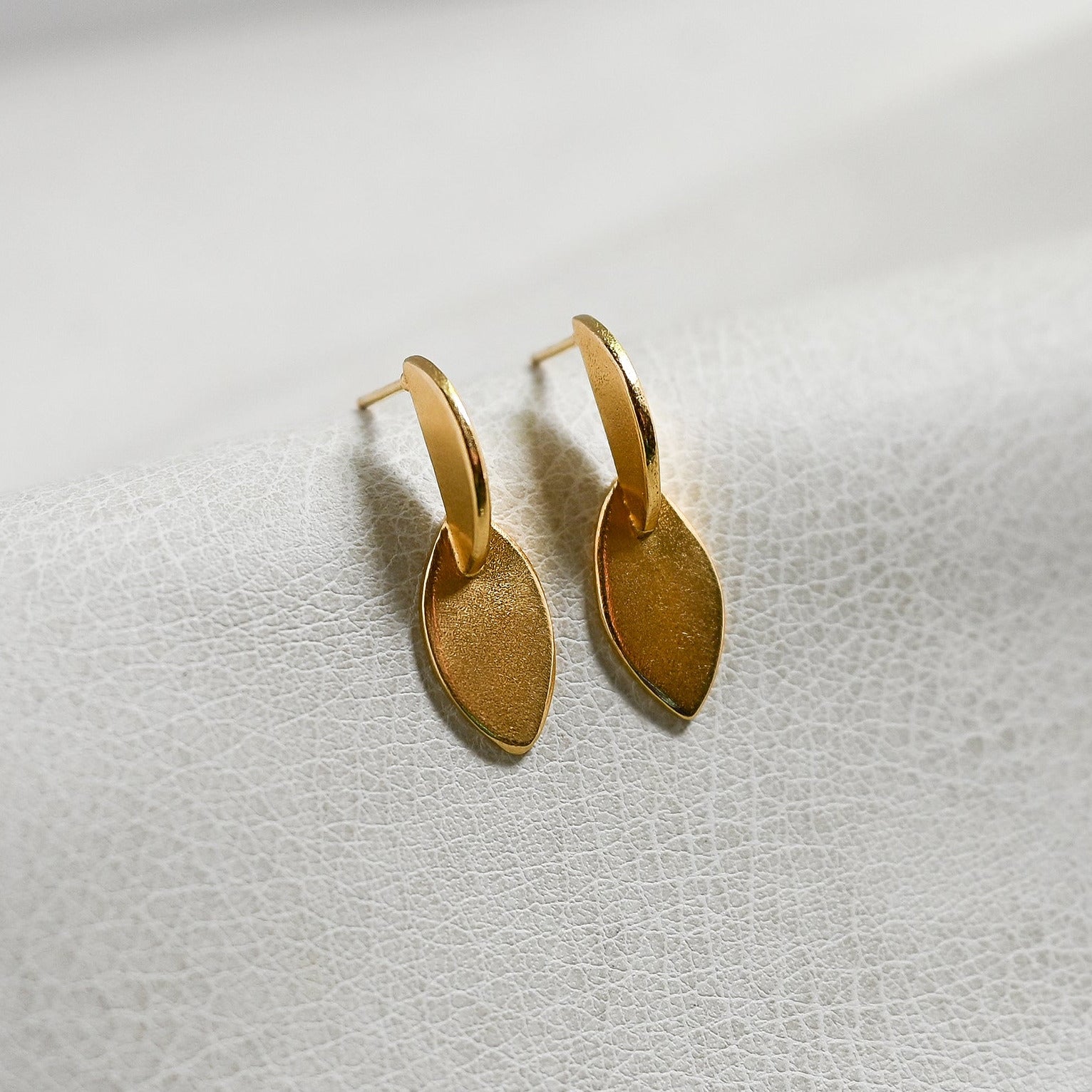 A pair of small gold leaf-shaped hoop earrings resting on a textured white surface.
