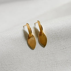 A pair of small gold leaf-shaped hoop earrings resting on a textured white surface.