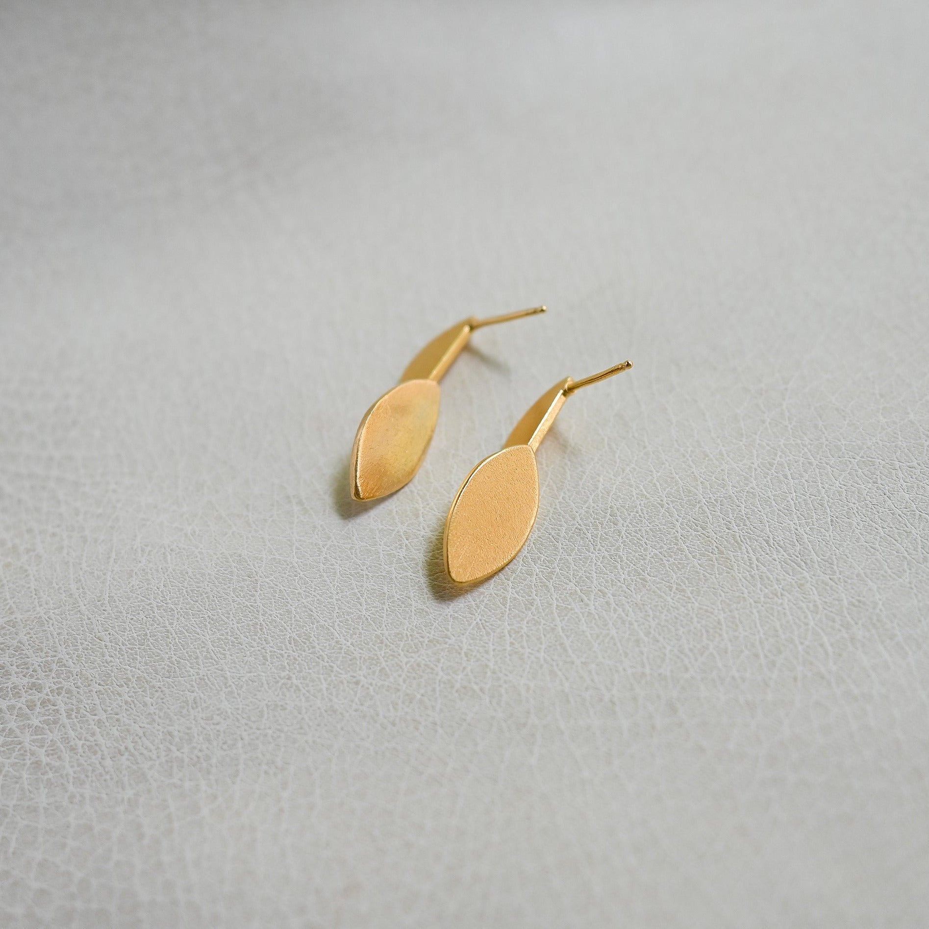 A pair of minimalist gold drop earrings with a flat, elongated oval pendant, displayed on a textured light gray surface.