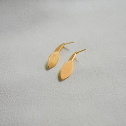 A pair of minimalist gold drop earrings with a flat, elongated oval pendant, displayed on a textured light gray surface.