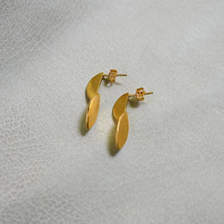 A pair of gold leaf-shaped stud earrings resting on a textured light gray surface.