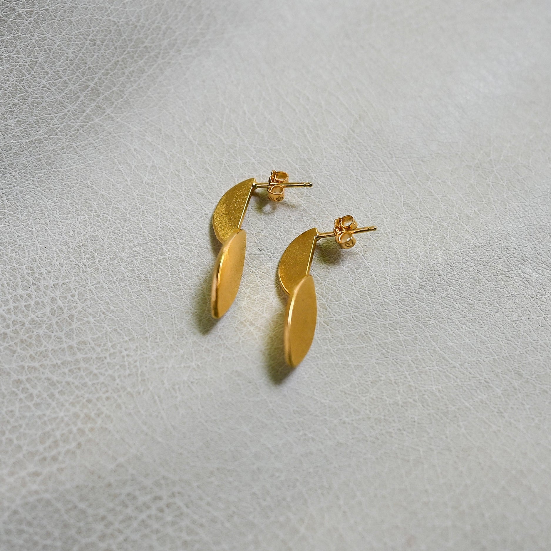 A pair of gold leaf-shaped stud earrings resting on a textured light gray surface.