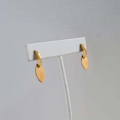 A pair of small gold leaf-shaped dangle earrings displayed on a white jewelry stand against a light gray background.