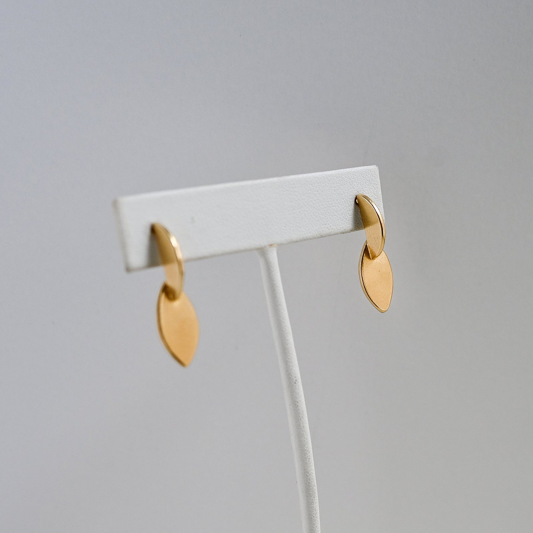A pair of small gold leaf-shaped dangle earrings displayed on a white jewelry stand against a light gray background.