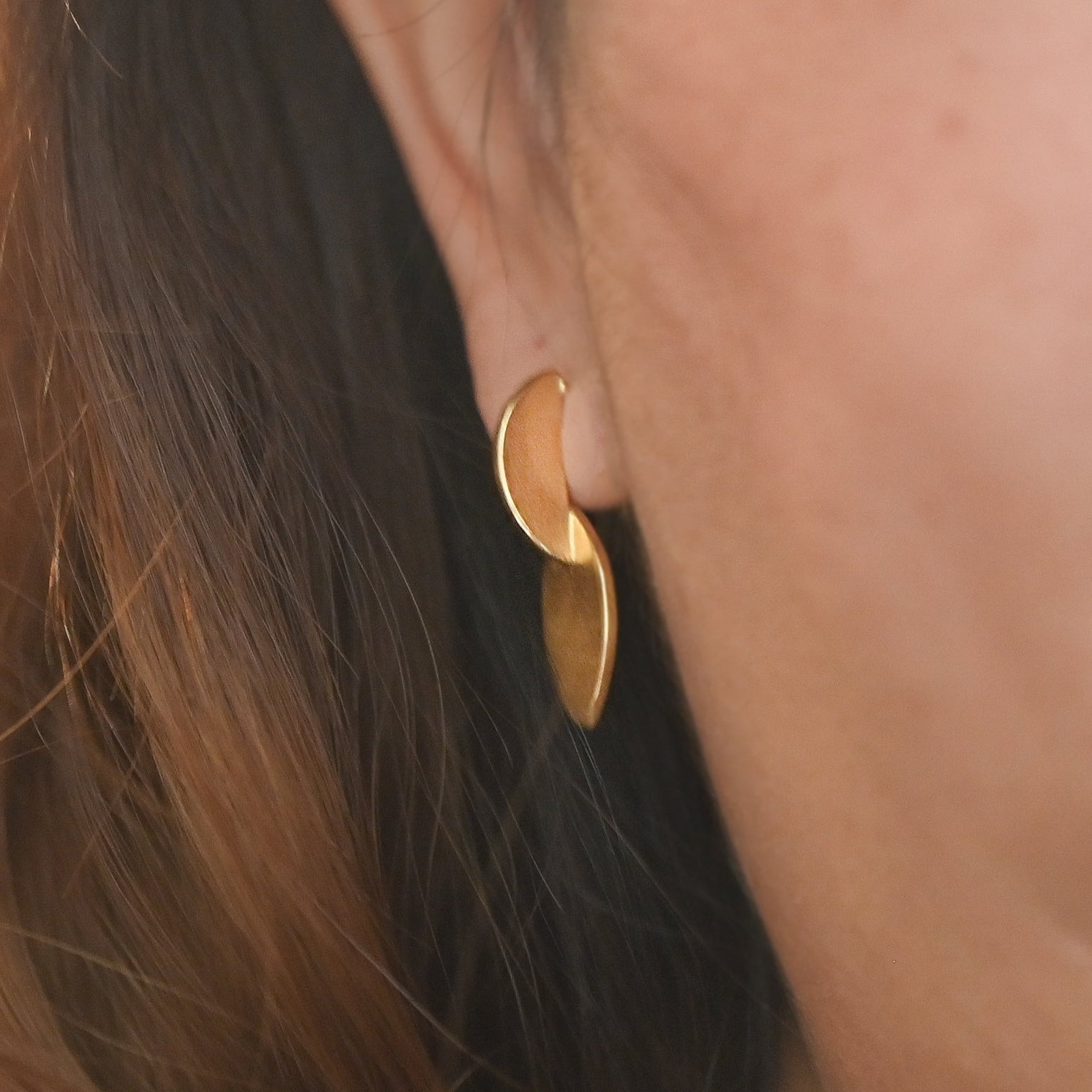 A close-up of a person's ear wearing a small gold crescent-shaped dangle earring, with dark brown hair and skin visible.