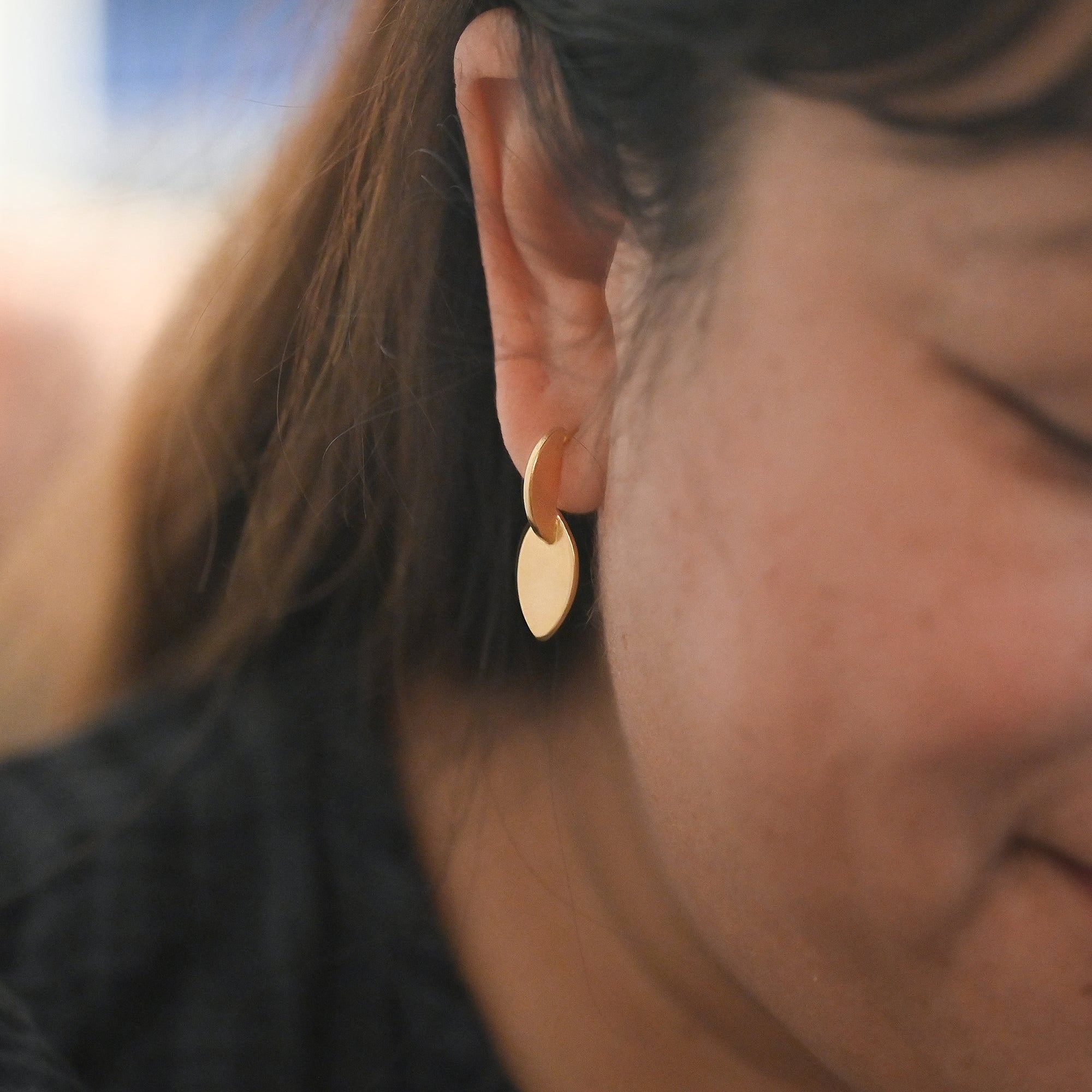 A close-up of a person's ear wearing a small gold hoop earring with a flat, disc-like gold drop pendant.
