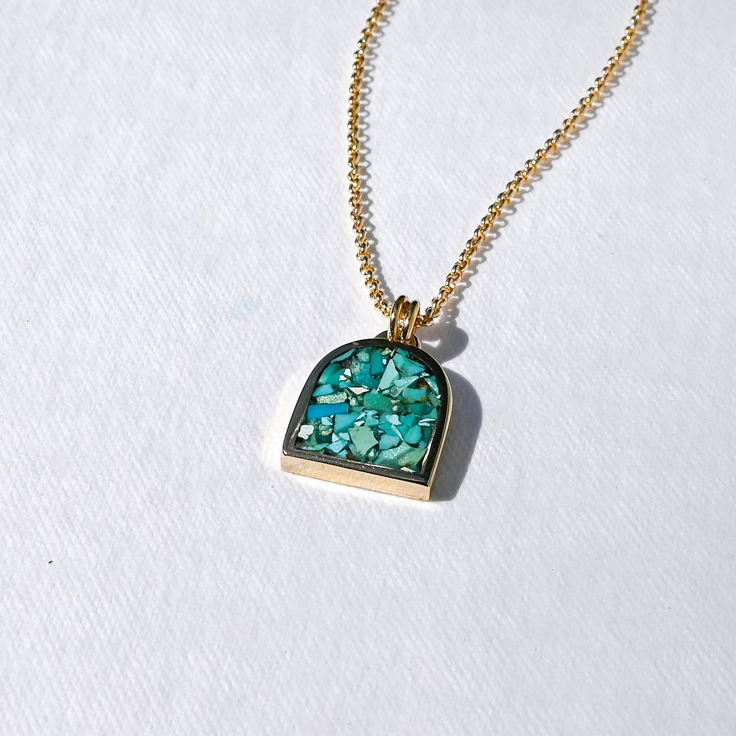 A gold chain necklace with a small arch-shaped pendant featuring a green and turquoise glittery inlay, displayed on a textured white surface.