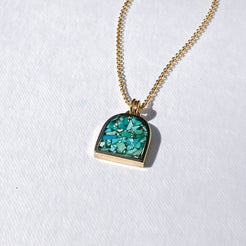 A gold chain necklace with a small arch-shaped pendant featuring a green and turquoise glittery inlay, displayed on a textured white surface.