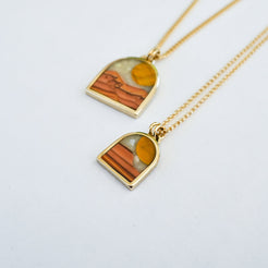 Gold necklace with a small arch-shaped pendant featuring a stone inlay design of tiger eye sun, orange and brown wave dolomite with a cream colored mother of pearl sky.