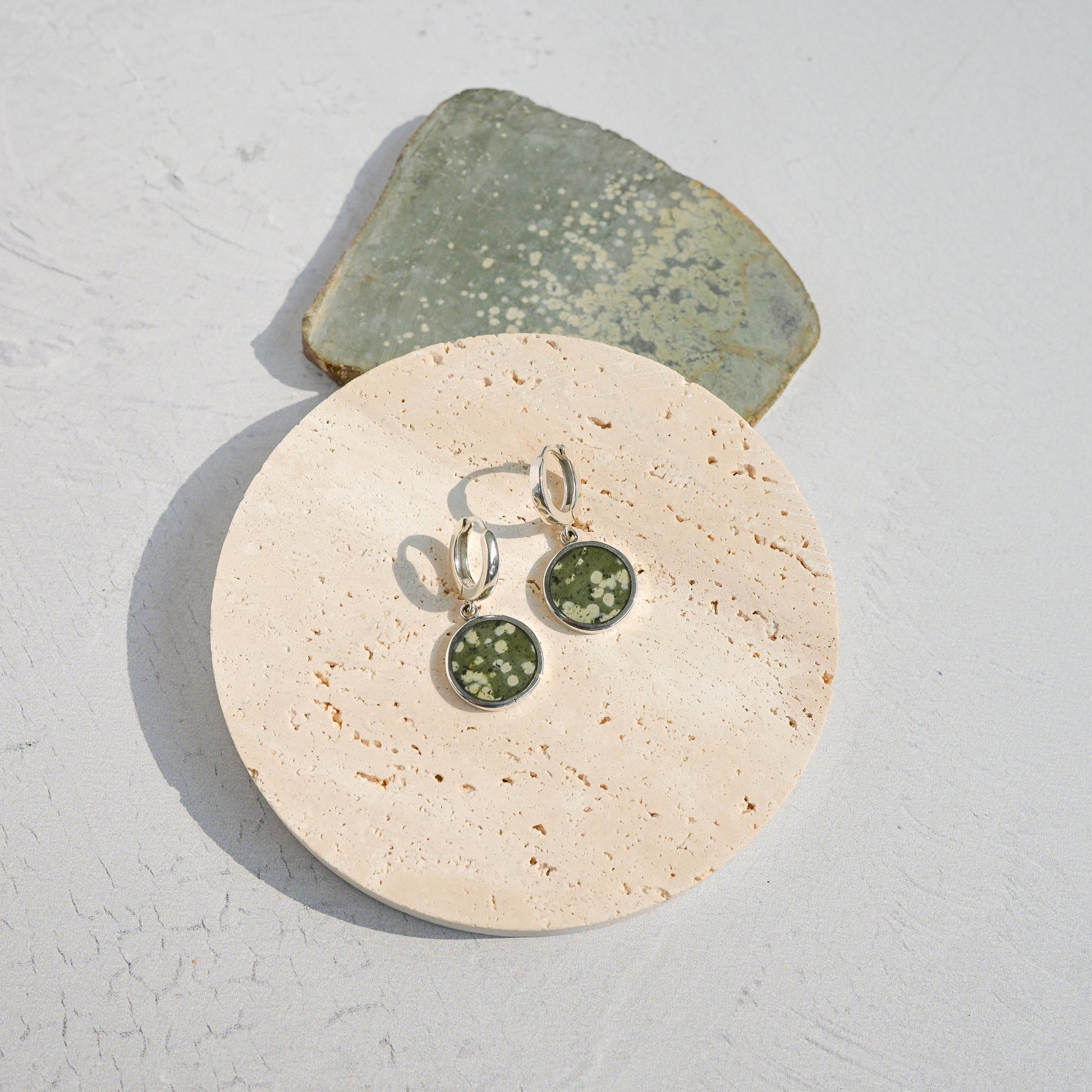 Pair of sterling silver huggie earrings with green variolite stone charms on a stone surface