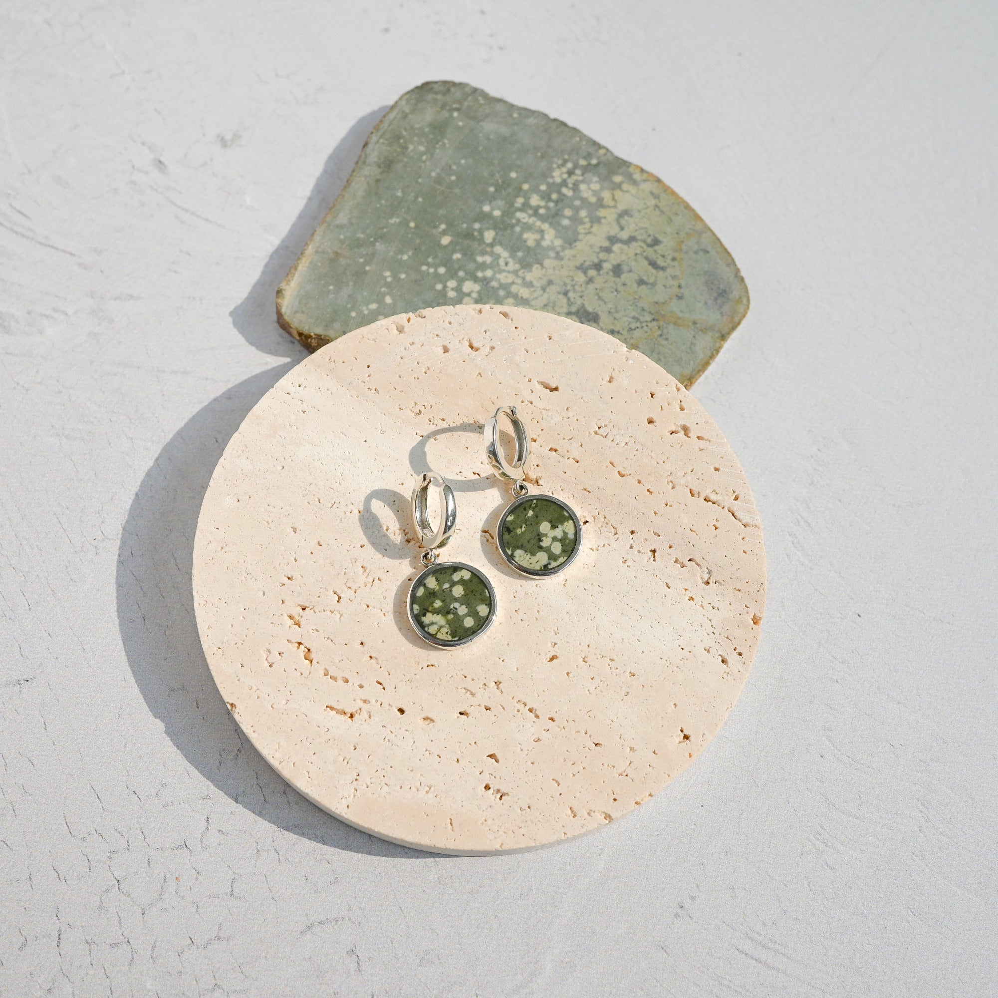 Pair of sterling silver huggie earrings with green variolite stone charms on a stone surface