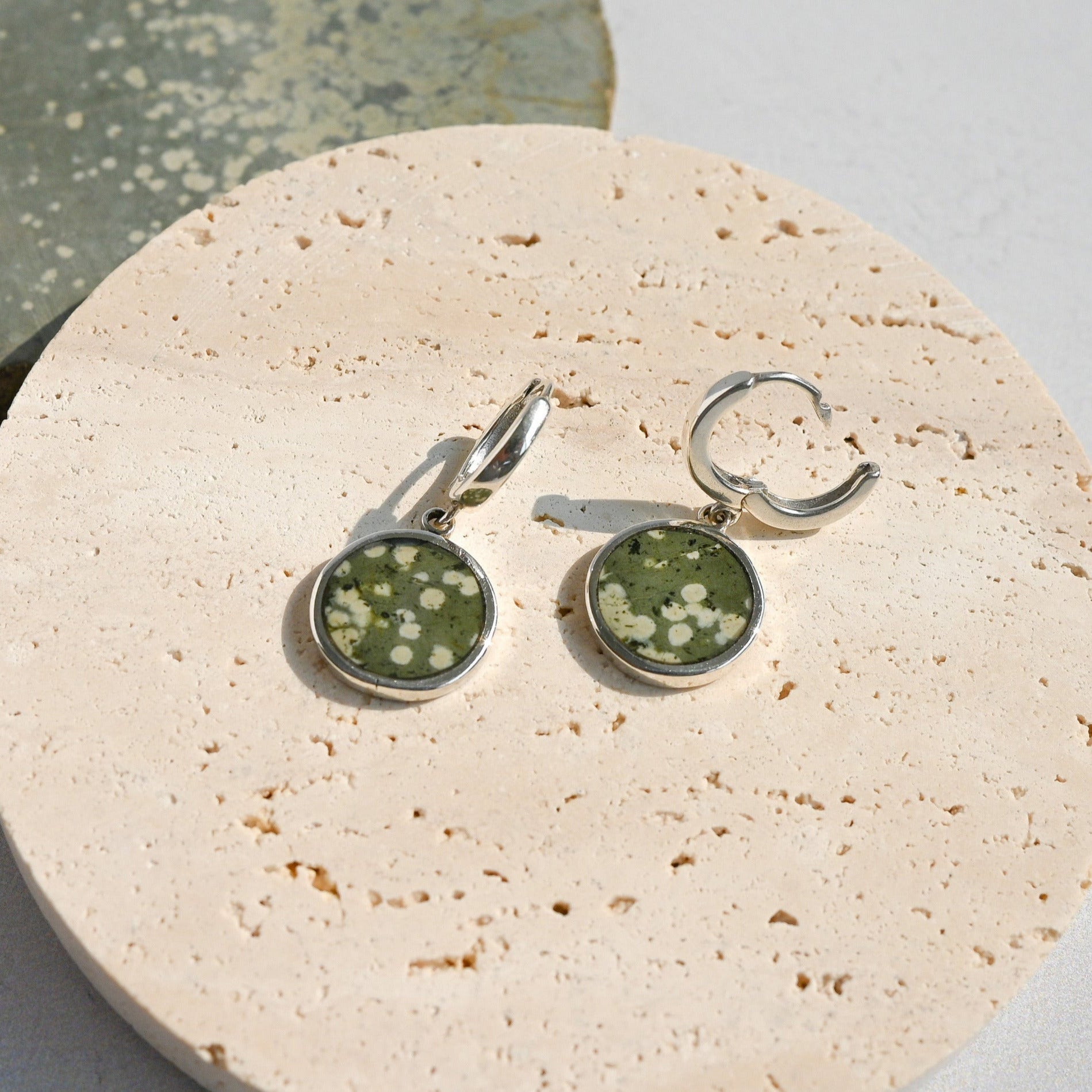 Pair of sterling silver huggie earrings with green variolite stone charms on a stone surface. One hoop is open.