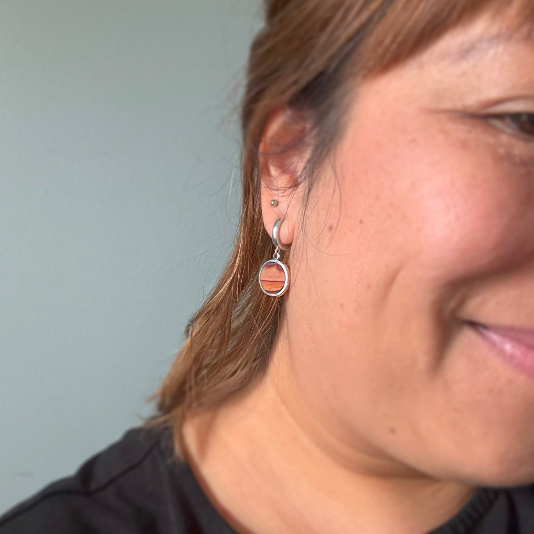 Close-up of a person wearing huggie hoop earring with dune-like stone charm on a round stone surface.