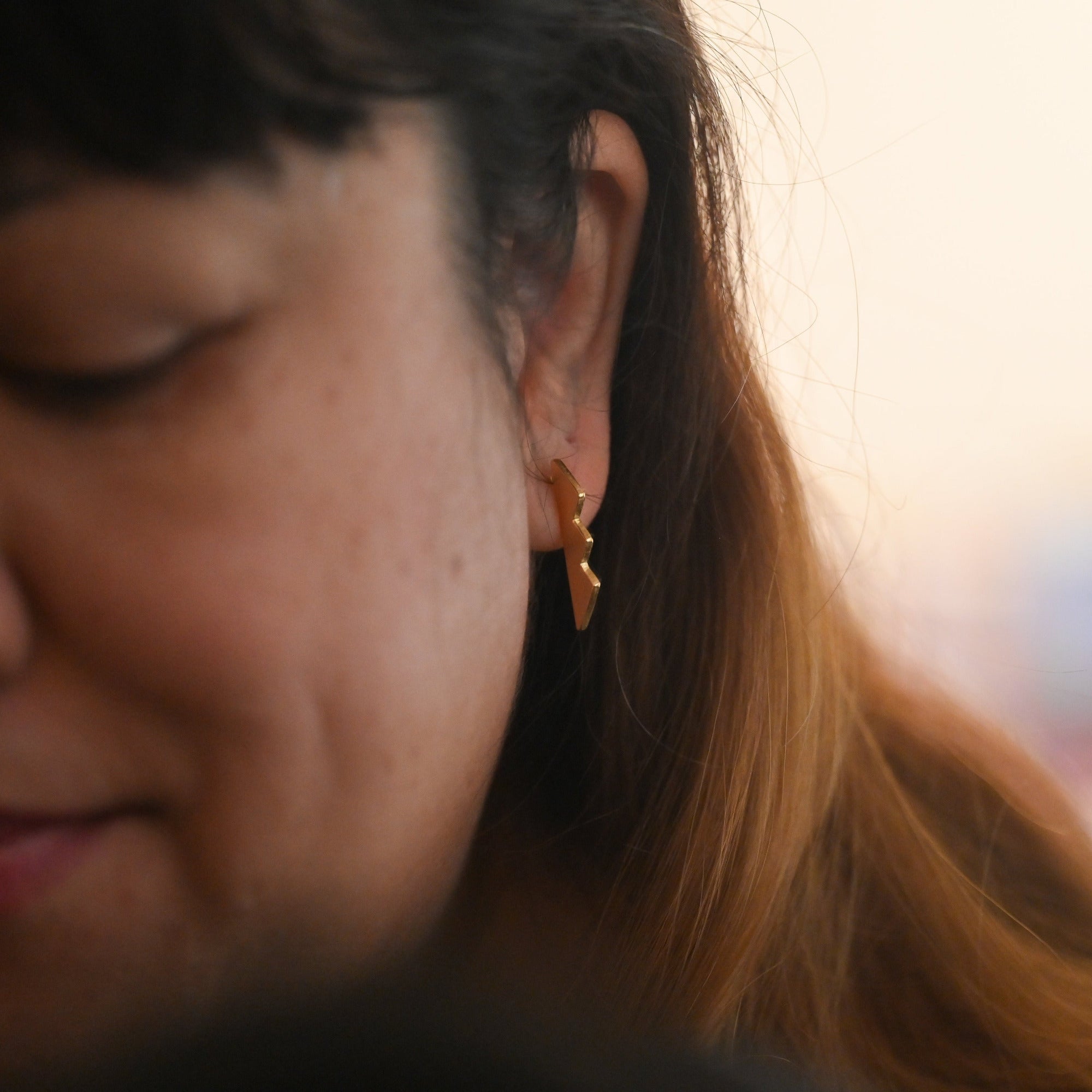 A close-up shot of a woman with dark hair wearing a small gold hoop earring, looking down with a soft, warm background.