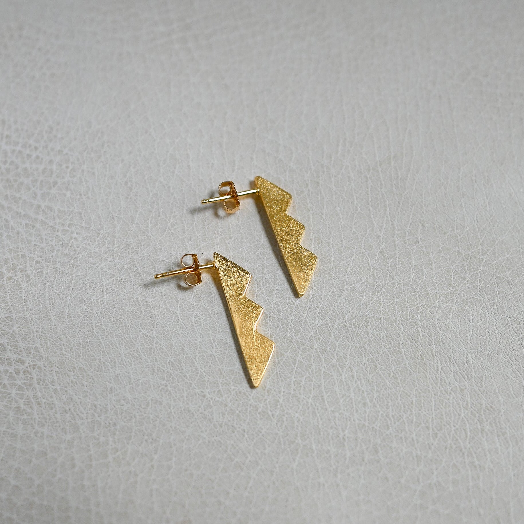 A pair of small gold lightning-bolt stud earrings on a light textured surface.