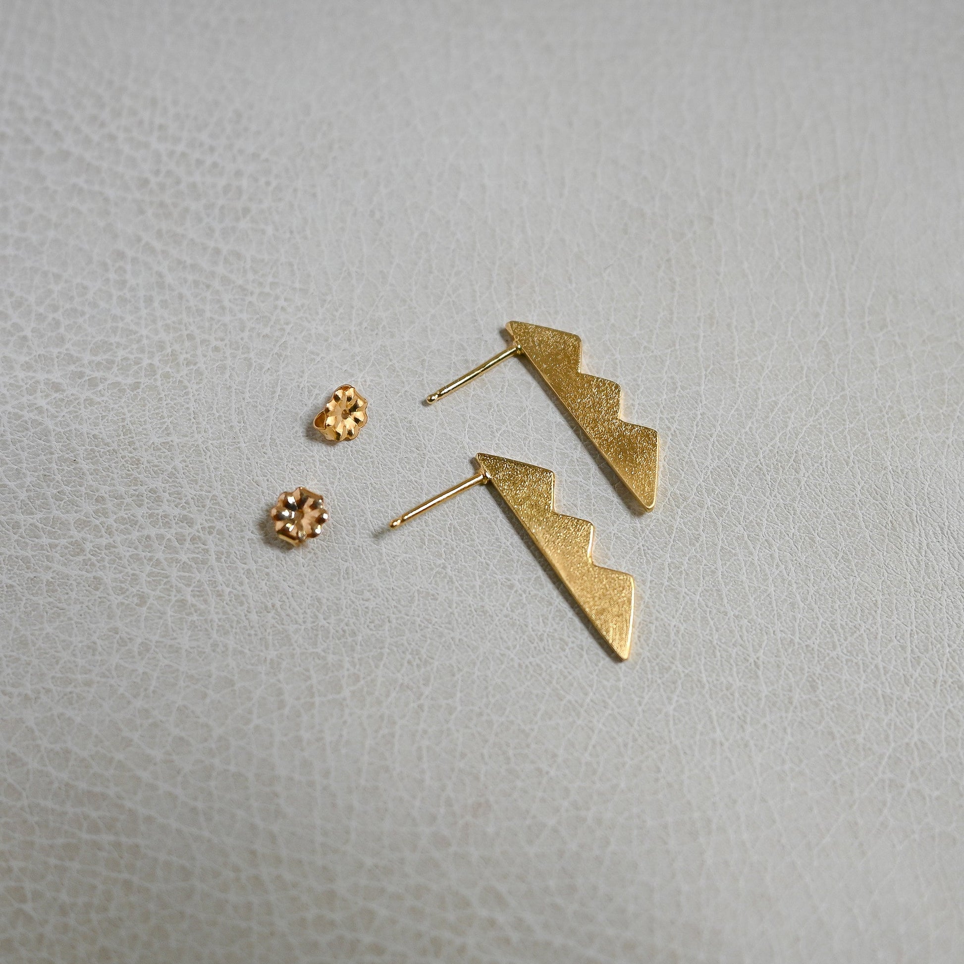 A pair of gold lightning bolt drop earrings with matching textured gold stud earrings, displayed on a light gray textured surface.