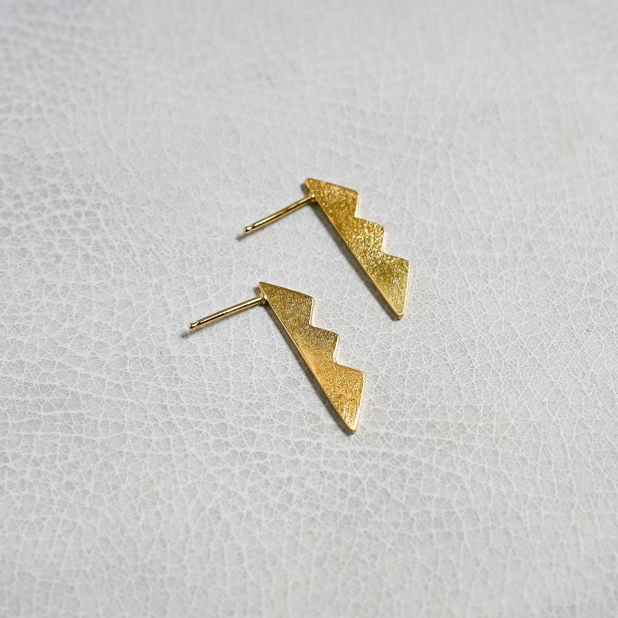 A pair of textured gold lightning-bolt stud earrings resting on a white textured surface.