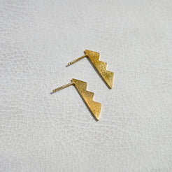 A pair of textured gold lightning-bolt stud earrings resting on a white textured surface.