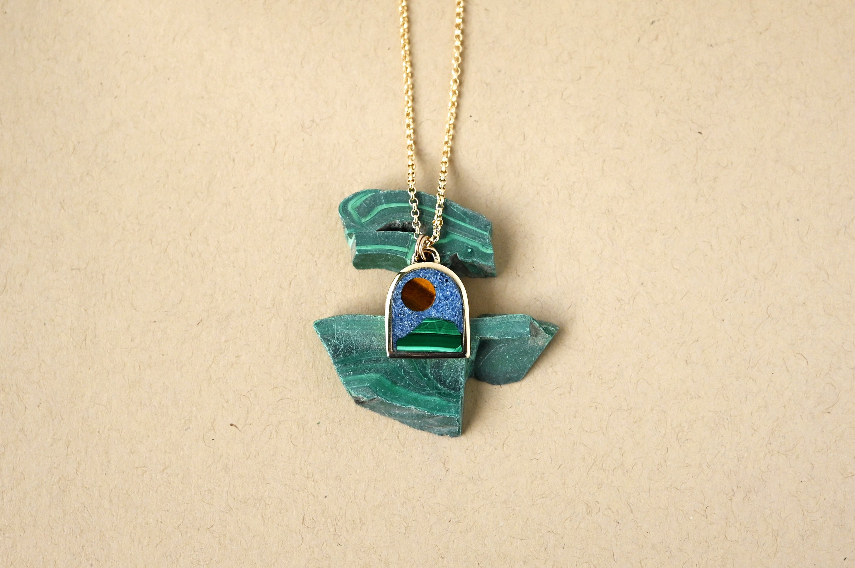 gold necklace with an arch shaped pendant featuring a scene made of  natural stones on a beige background