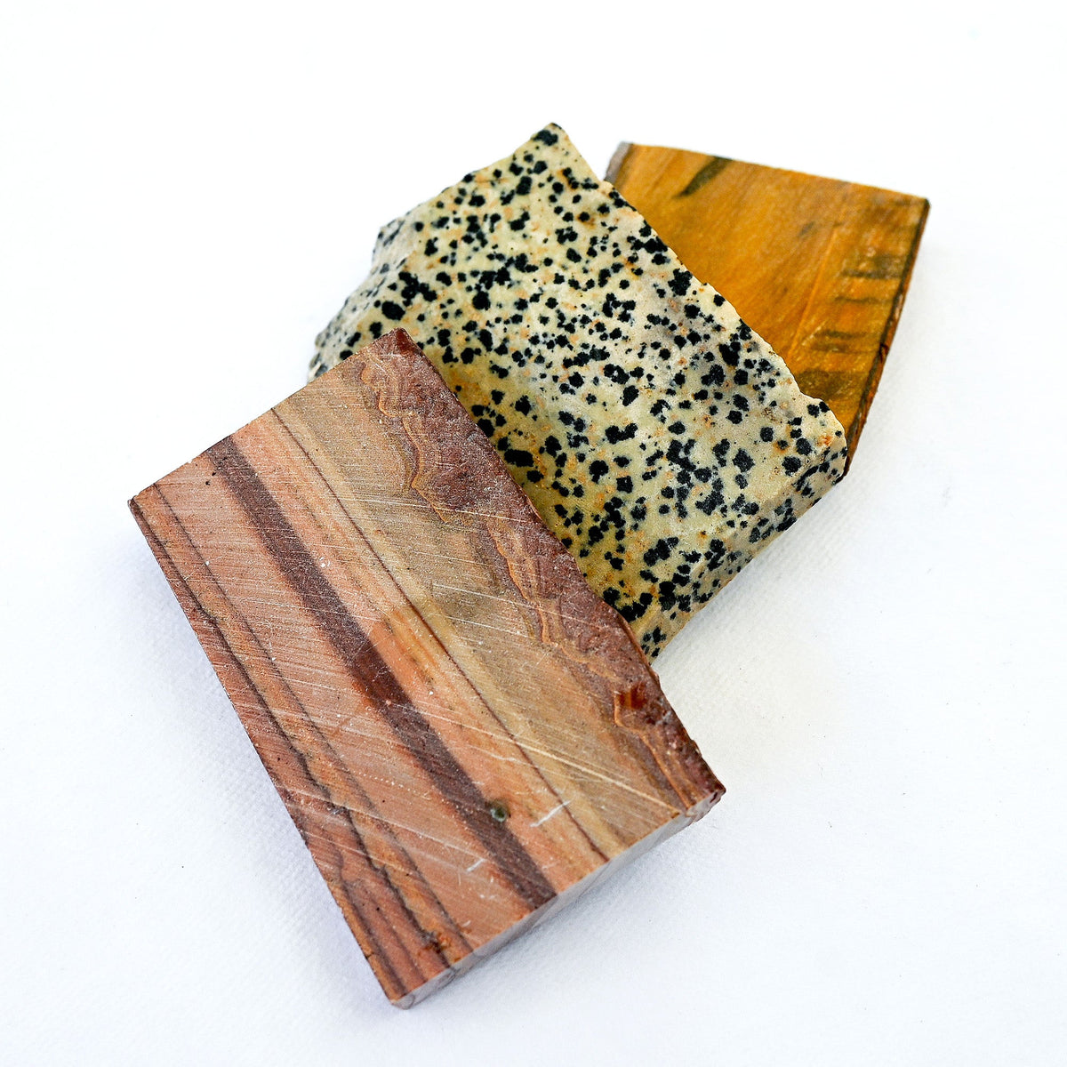 Three rectangular stone or wood blocks with distinct textures and colors, including a speckled beige and black stone, a reddish-brown wood grain, and a golden-brown wood grain, arranged on a plain white background.